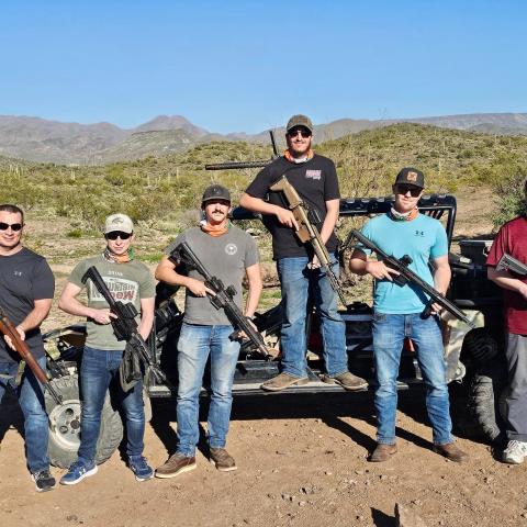 Six men holding rifles stand in front of a desert landscape with mountains.