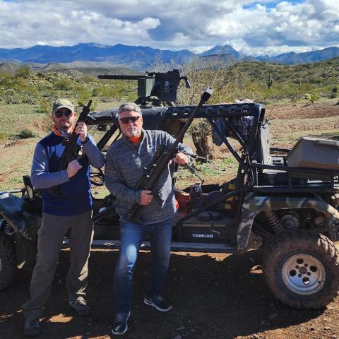 Two men holding rifles stand by an off-road vehicle with mountains in the background.