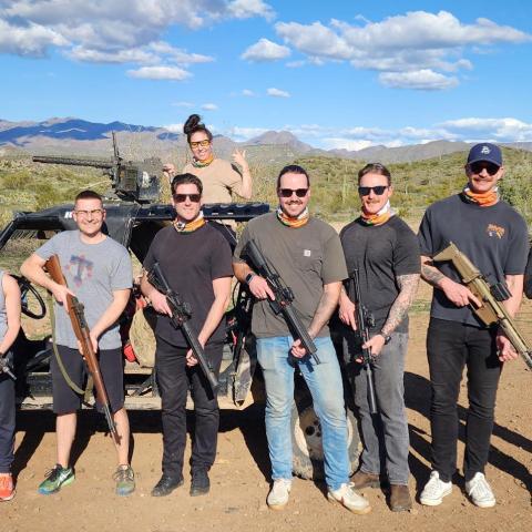 Group of nine people with large rifles standing outdoors in a desert landscape with mountains in the background.