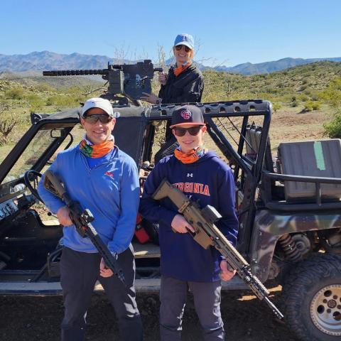 Three people outdoors with rifles beside an off-road vehicle in a desert landscape.