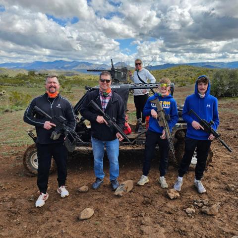 Five people holding rifles pose outdoors near a small off-road vehicle, with mountains in the background.