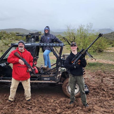 Three people with rifles stand by a rugged buggy in a muddy desert landscape.