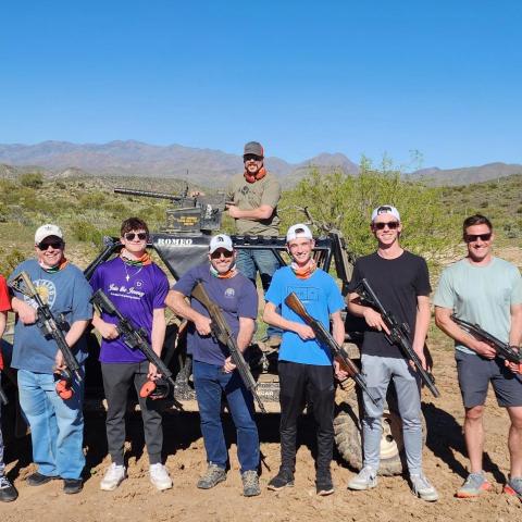 Group of nine people holding paintball guns in a desert landscape with a vehicle in the background.