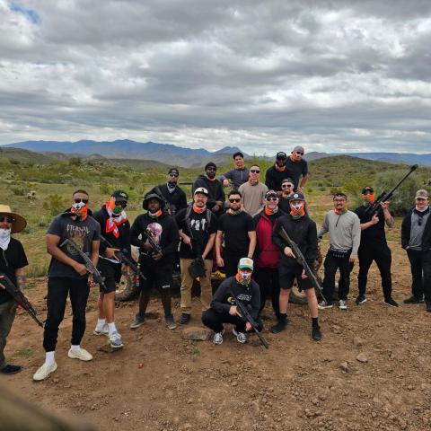 Group of people outdoors, some holding rifles, wearing casual clothing, standing on rocky terrain.