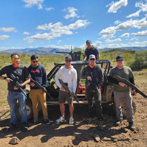 Six people with firearms pose in front of an off-road vehicle in a desert landscape.