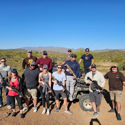 Group of people posing with rifles in a desert landscape, with cacti and mountains in the background.
