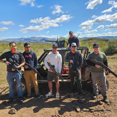 Six people in masks holding rifles, standing in front of an off-road vehicle in a desert landscape.