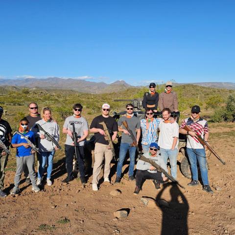 Group of people posing with rifles outdoors in a desert landscape.