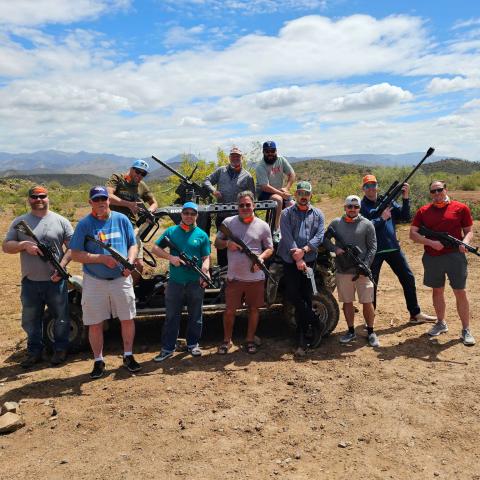 Group of men with rifles standing around an off-road vehicle in a desert landscape.