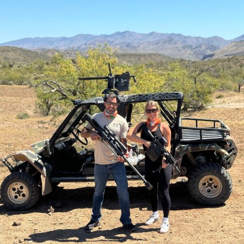 Two people with rifles stand in front of an ATV in a desert landscape.