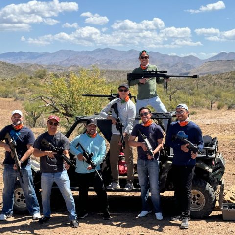 Group of seven people holding rifles poses near a vehicle in a desert landscape under a blue sky.