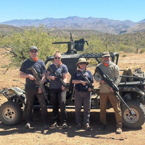 Four people with rifles stand in front of an off-road vehicle in a desert landscape.
