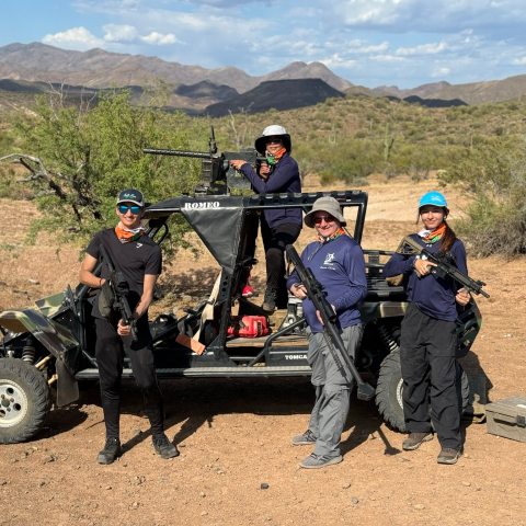 Four people with guns posing by an off-road vehicle in a desert landscape.