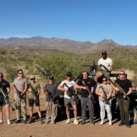 Group of people posing with firearms in a desert landscape with mountains in the background.