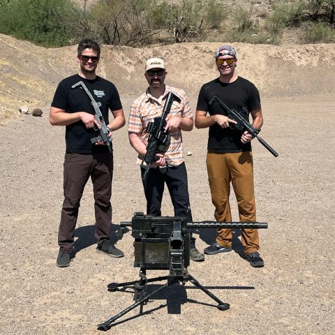 Three people outdoors holding guns, positioned behind a large mounted weapon on a sunny day.