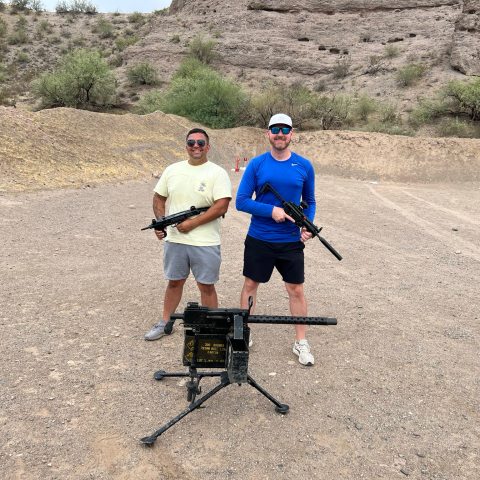 Two people holding rifles at a shooting range with desert landscape in the background.
