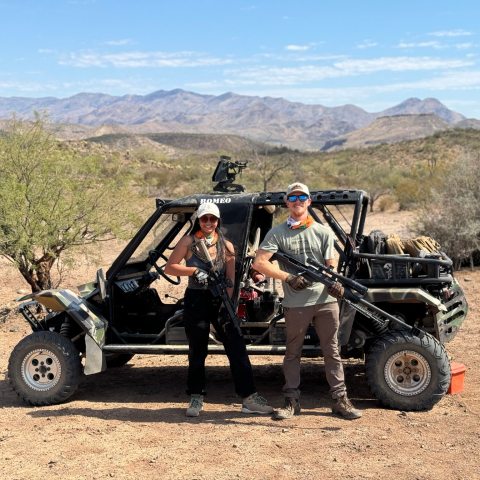 Two people with rifles stand by an off-road vehicle in a desert landscape with mountains in the background.