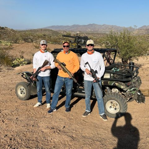 Three men with rifles stand beside a camouflaged ATV in a desert landscape.