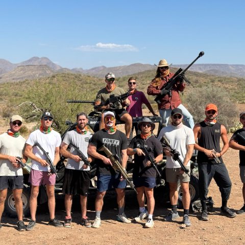 Group of people posing with rifles in a desert landscape, standing and sitting on a vehicle.