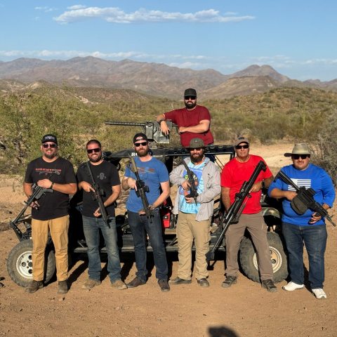 Seven men with firearms stand in front of an off-road vehicle in a desert landscape.