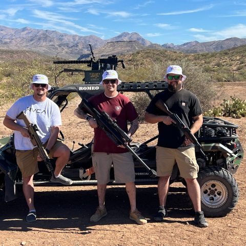 Three men holding rifles stand by an off-road vehicle in a desert landscape.