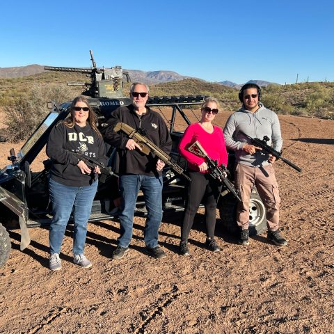 Four people with firearms stand by an off-road vehicle in a desert landscape.