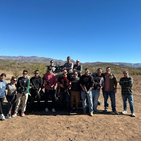 Group of 14 people posing outside with a desert landscape and mountains in the background.