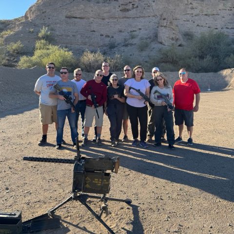 Group of people posing with rifles on a dirt range with a machine gun in front. Rocky terrain in the background.