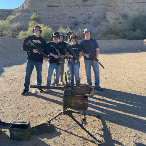 Group of five people holding rifles standing outdoors near desert cliff.