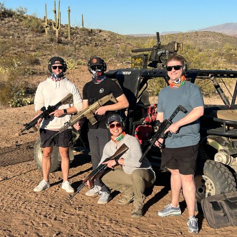 Four people with rifles and masks pose by a vehicle in a desert landscape.