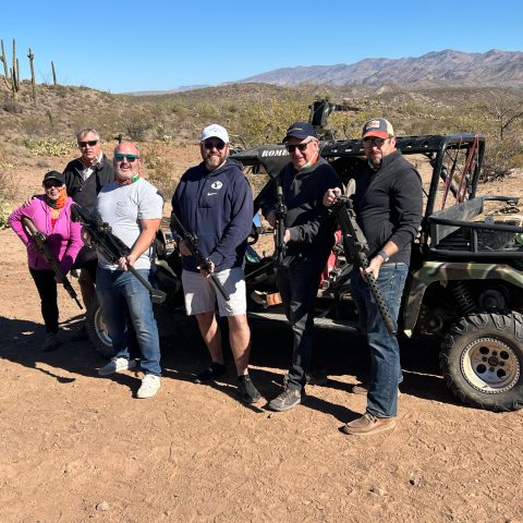 Group of people holding rifles stand in front of an off-road vehicle in a desert landscape.