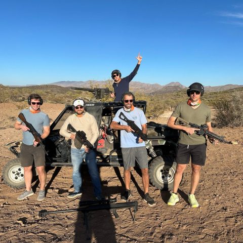 Five people with firearms pose in front of an off-road vehicle in a desert landscape.