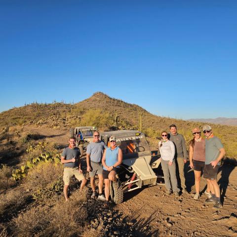 Group posing by off-road vehicle in desert landscape under clear blue sky.