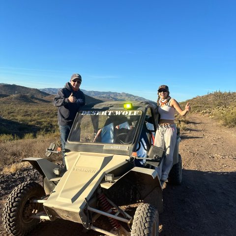 Two people posing with a desert buggy on a dirt path under a clear blue sky.