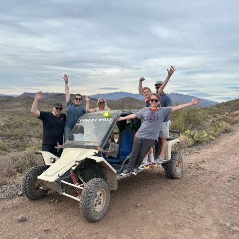 Six people posing with a dune buggy in a desert landscape, raising their arms and smiling.