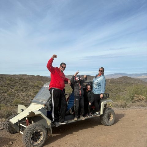 Four people celebrating atop an off-road vehicle in a desert landscape.