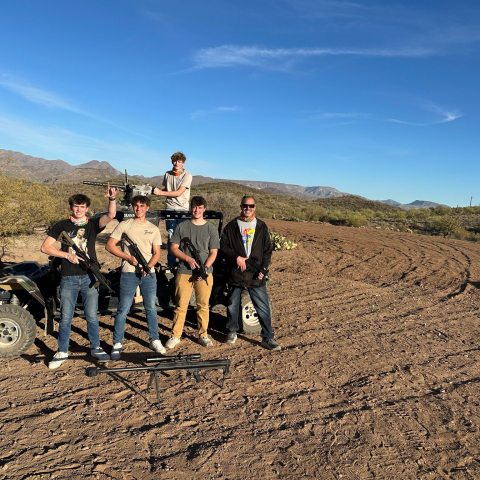 Group of five people outdoors holding rifles, standing near an ATV in a desert landscape.