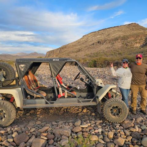 Two people stand beside an off-road vehicle on rocky terrain with hills in the background.