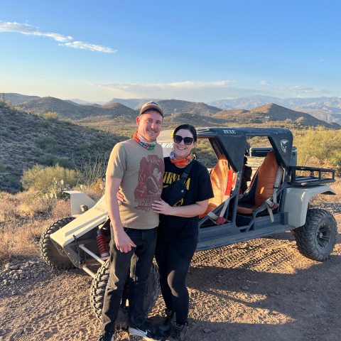 Couple wearing bandanas standing by an off-road vehicle in a desert landscape.