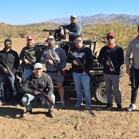 Group of eight people with rifles posing near an off-road vehicle in a desert landscape.