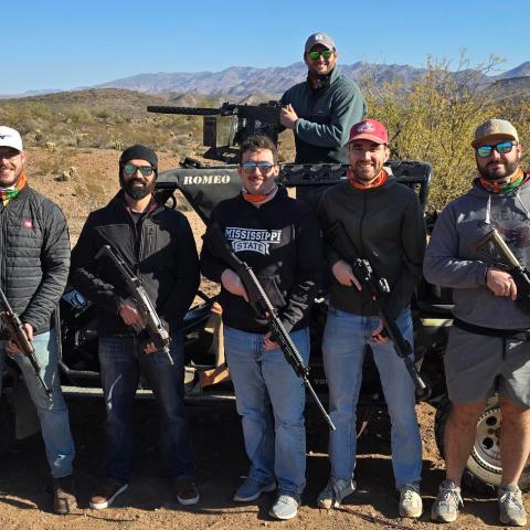 Group of six men with rifles in a desert, standing by an off-road vehicle.