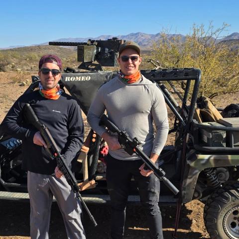 Two people with rifles stand beside an off-road vehicle in a desert landscape.
