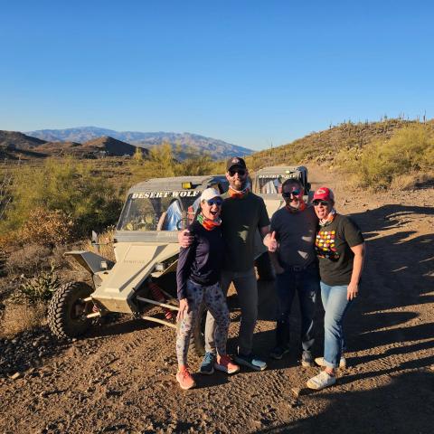 Four people standing in front of off-road vehicles in a desert landscape under clear blue skies.