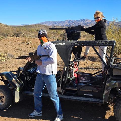 Two people on a dune buggy in desert with rifles, cacti and mountains in background.