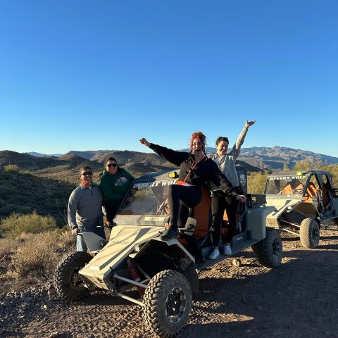 Four people posing with an off-road vehicle on a dirt trail against a mountain backdrop.