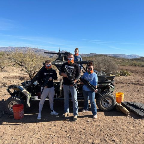 Group of people with masks and rifles in front of an off-road vehicle in a desert landscape.
