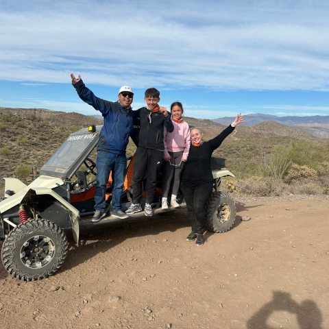 Four people stand on an off-road vehicle on a desert trail with mountains in the background.