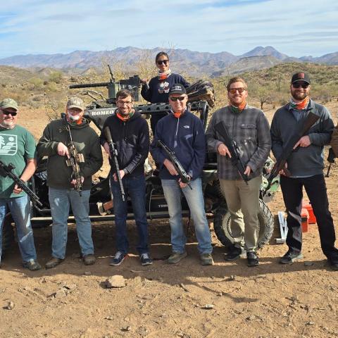Eight men with rifles pose in front of an off-road vehicle in a desert setting.