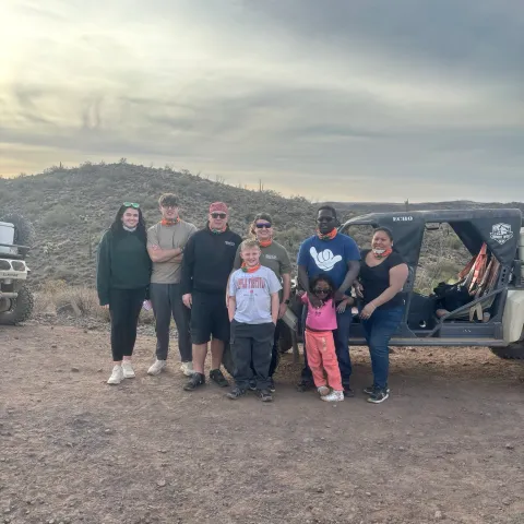 Group of people posing together in desert landscape near a vehicle, under a cloudy sky.