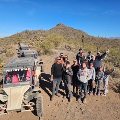 Group of people posing in desert with off-road vehicles behind.